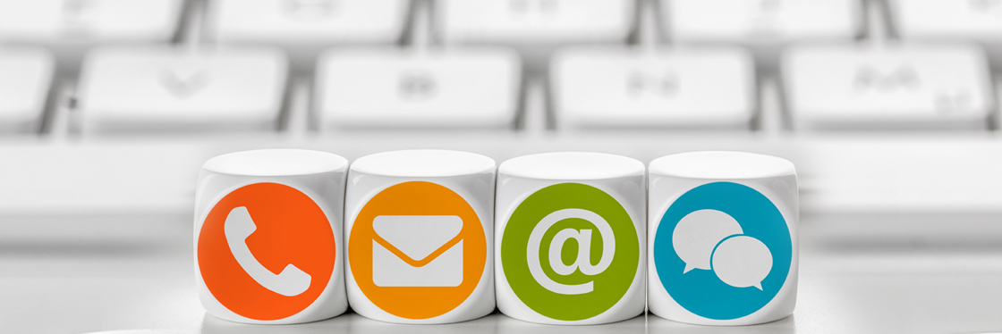 A keyboard in the background, with four dice lined up in front. The dice each have a symbol on them: a red circle with a phone, an orange circle with an envelope, a green circle with a @, and a blue circle with two text bubbles.