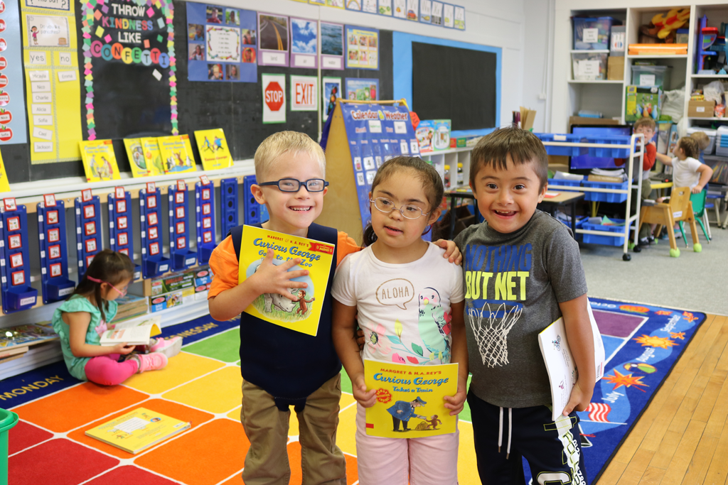 Three children stand together, all holding Curious George books. They are in a school classroom.