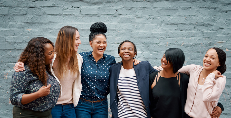 Six women gathered together with their arms around each other, smiling and laughing. A gray brick wall is behind them.