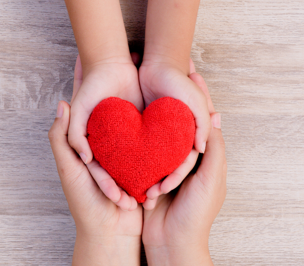 An adult's hands cradling a child's hands, which are cradling a red knitted heart.