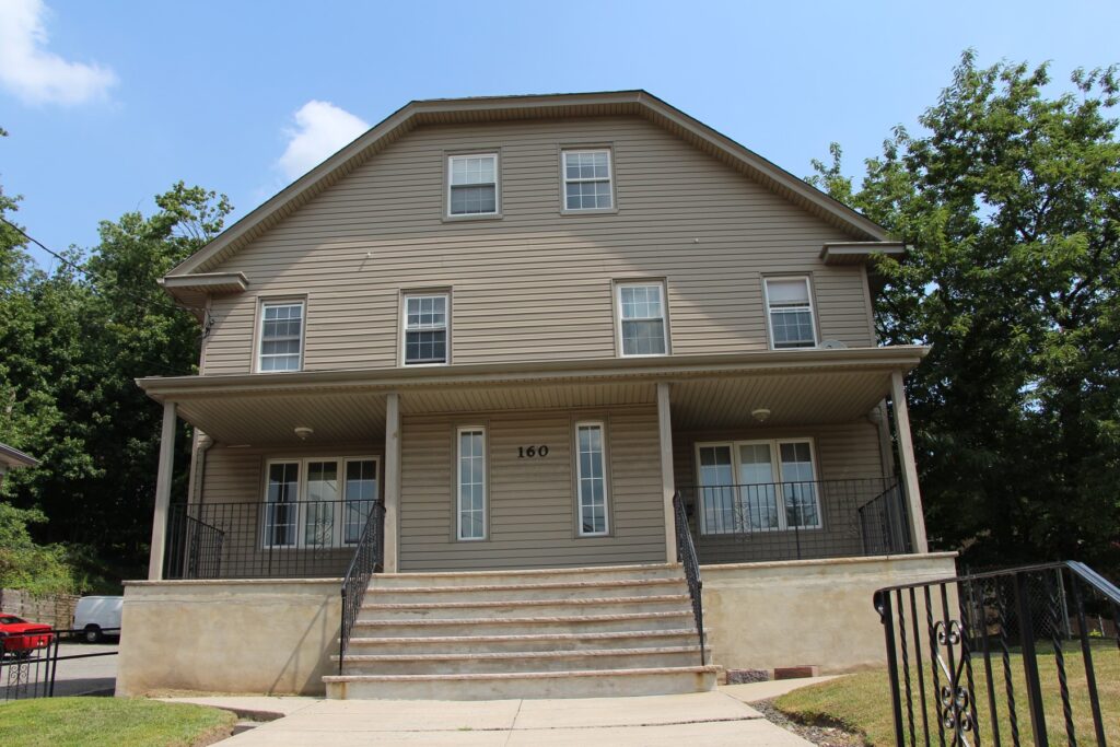 A photo of the front of a home, with steps leading up to it. The home is a tan color and includes porch areas that have wrought-iron railings.