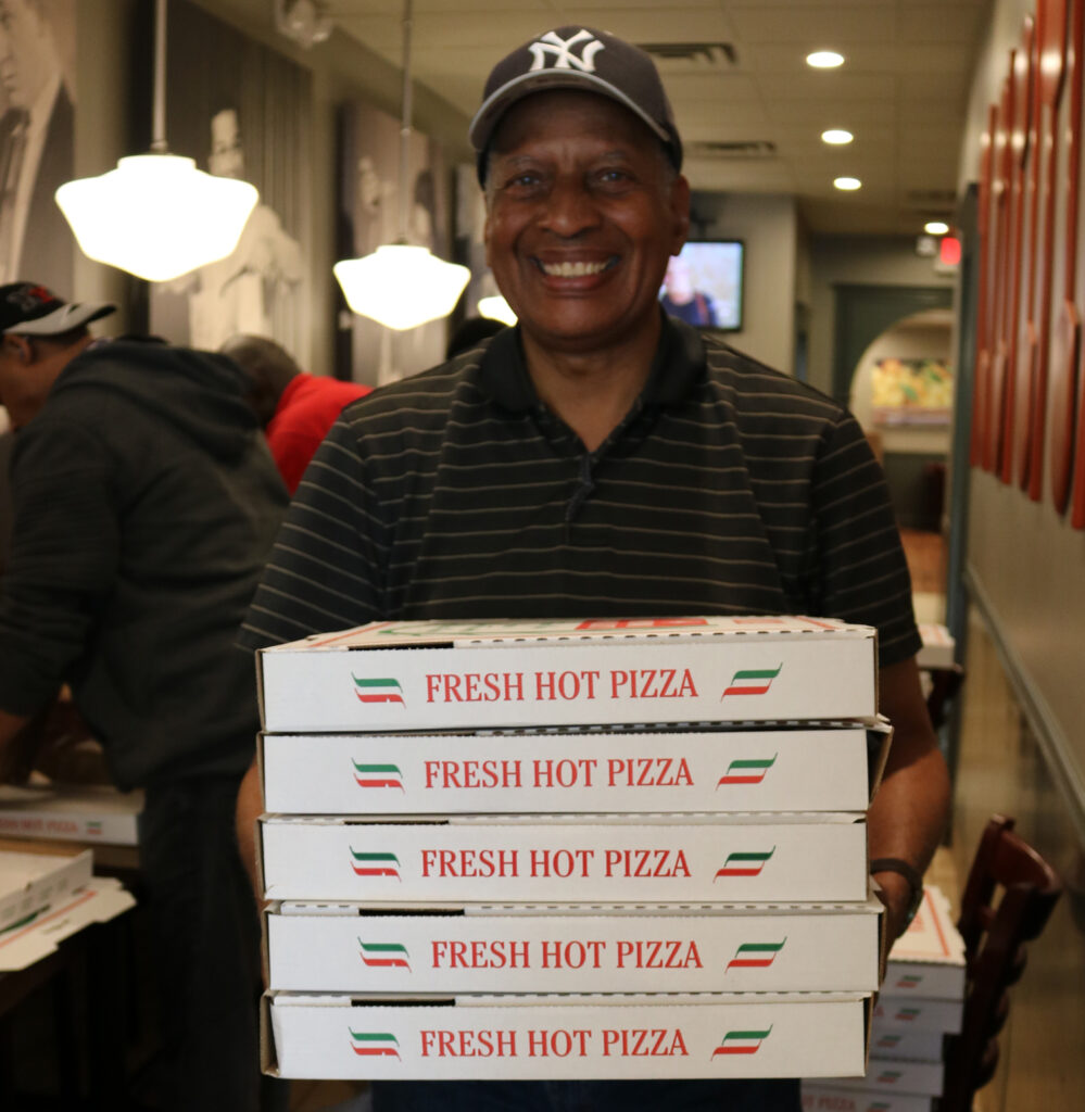 A man standing in a restaurant, smiling, while holding a stack of pizza boxes in front of him.