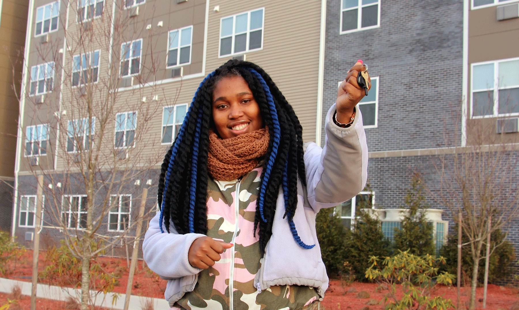 A woman standing in front of an apartment building, holding keys in her hand.