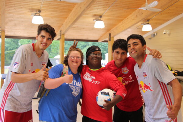 Five people gathered together under a pavilion, wearing Red Bulls gear. The boy in the center is holding a soccer ball.