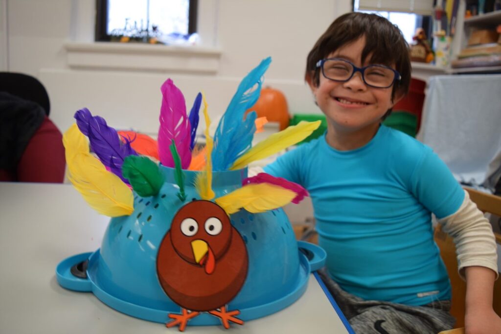 A boy in glasses and a blue shirt sitting next to a school table, where there is a turkey craft of feathers sticking out of a blue colander.