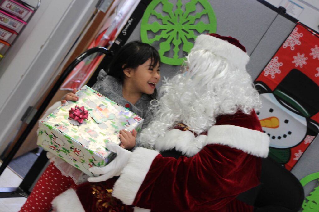 A girl holding a wrapped holiday gift and smiling and laughing while she's looking at Santa. Snowman and snowflake decorations are visible in the background.