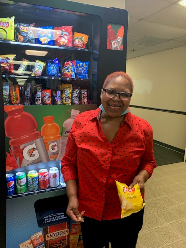 A woman in a red shirt standing in front of a vending machine, holding a bag of potato chips.