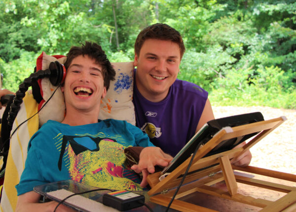 Two people sitting together at Camp Hope, using an electronic devise positioned on an easel. Both are smiling. The boy on the left is in an electric wheelchair.